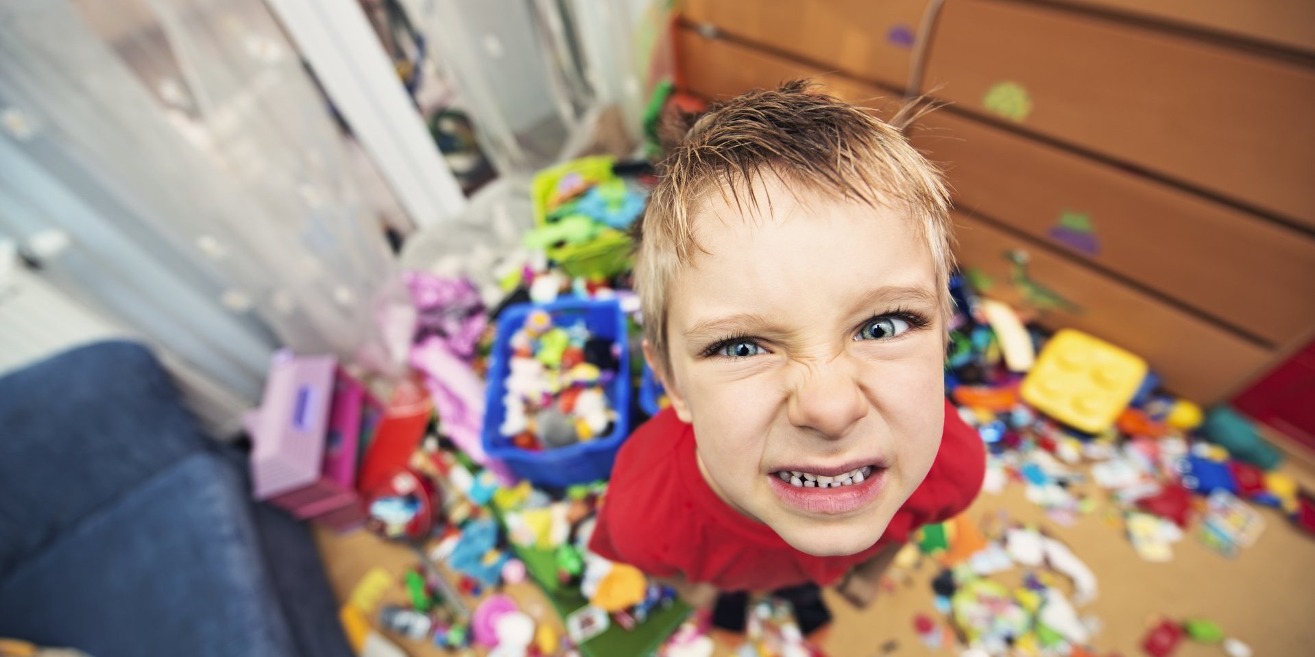 Kind steht in einem unaufgeräumten Zimmer zwischen verstreutem Spielzeug und zieht eine angespannte Grimasse