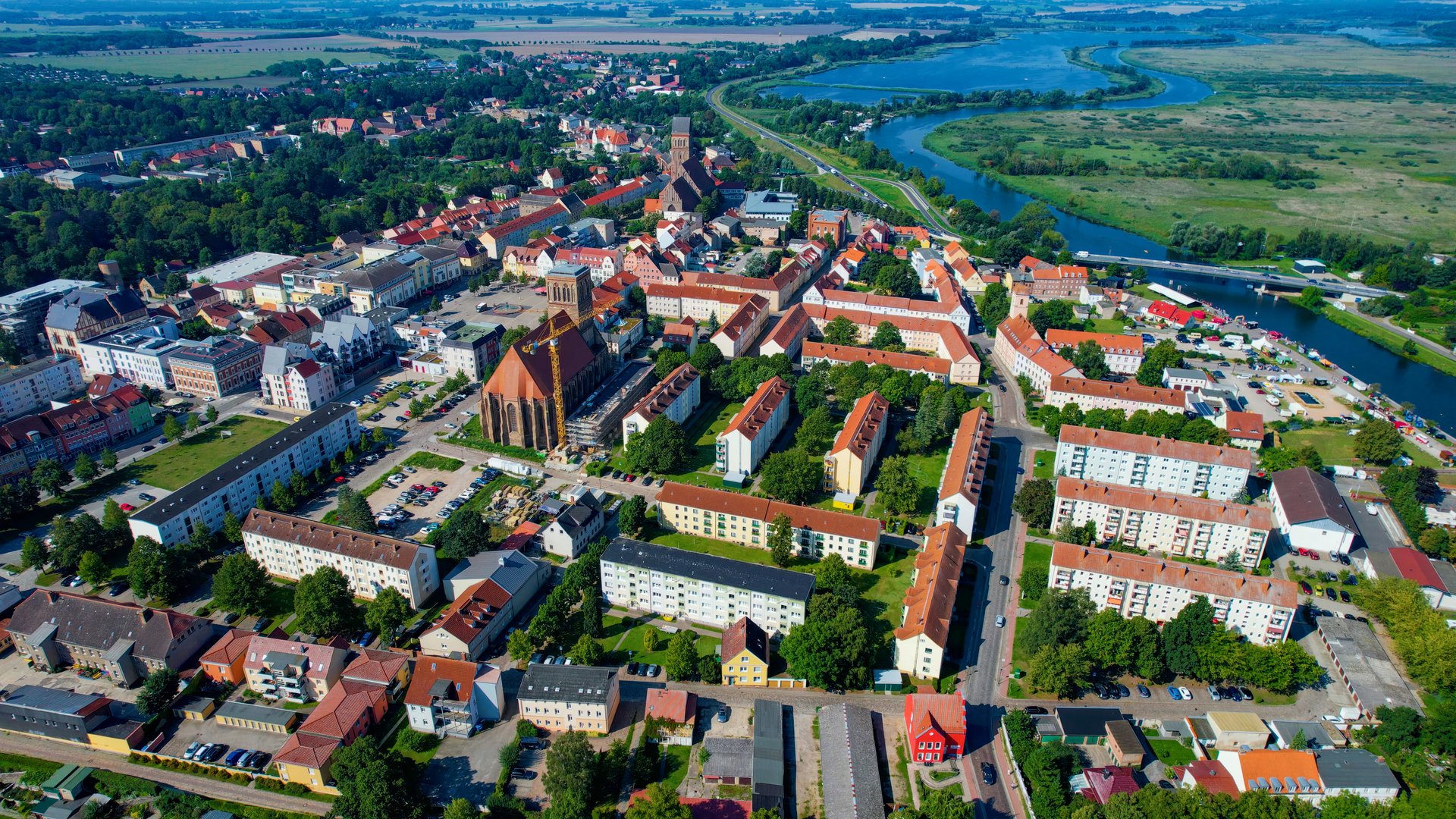 Luftaufnahme einer Stadt mit Wohngebieten, Kirchturm und umliegender Seenlandschaft