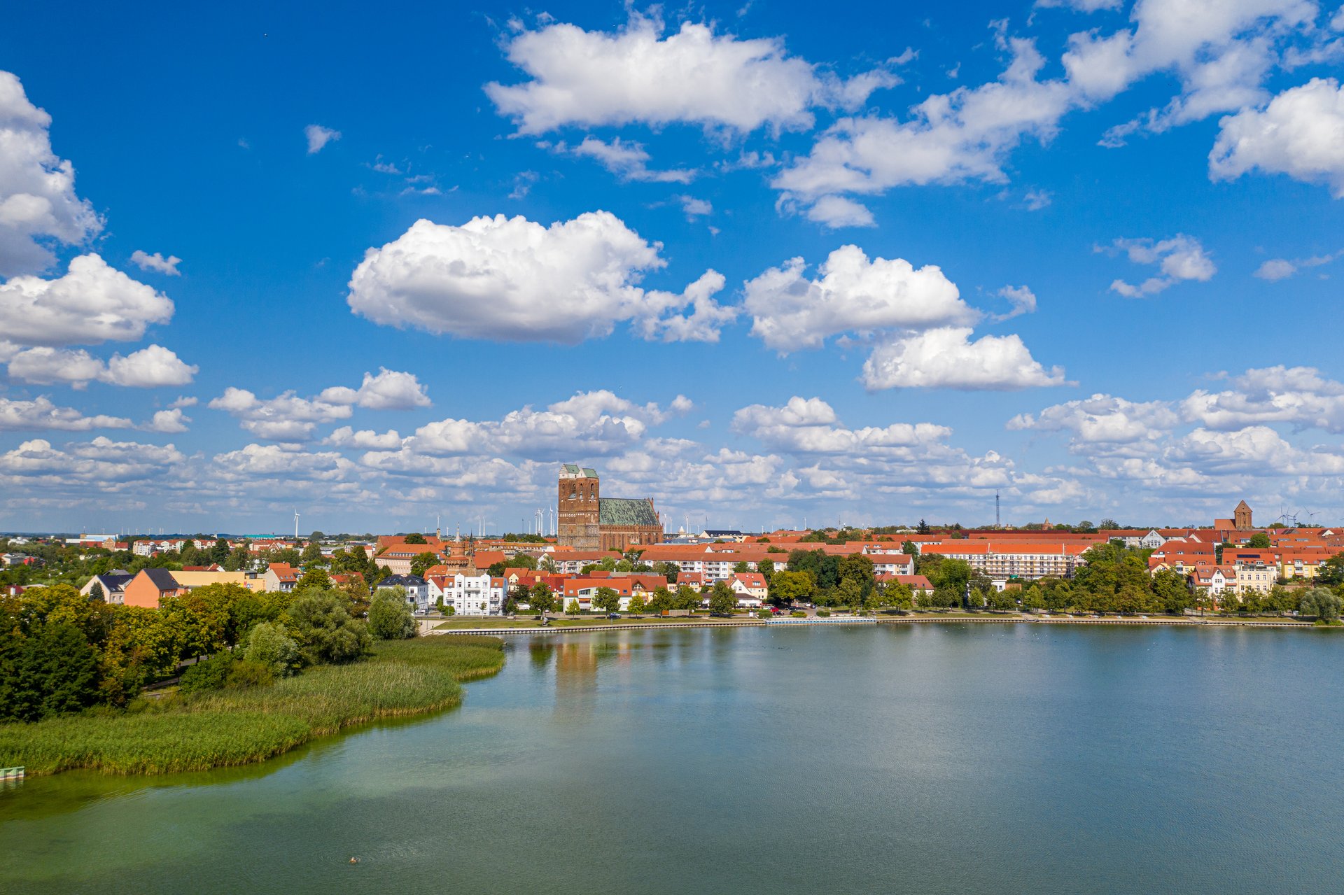 Blick über einen See auf eine Stadt mit Kirchturm unter blauem Himmel.